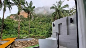 A modern bathroom with a large white bathtub, a stone wall, and a view of a lush green forest. - Red Cheek Mountain Villa  (Krabi)