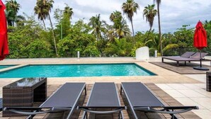 A swimming pool with lounge chairs and red umbrellas, surrounded by palm trees and greenery. - Feel at home in paradise/ A few minutes from the beach. (Nuevo Vallarta)