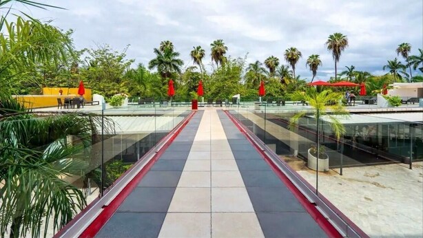 A glass bridge with a red railing, surrounded by palm trees and greenery, leading to a building with red umbrellas. - Feel at home in paradise/ A few minutes from the beach. (Nuevo Vallarta)
