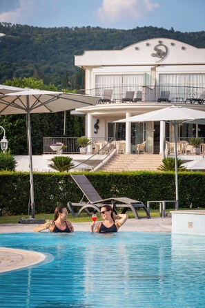 Ein Schwimmbecken mit zwei Personen, ein Gebäude mit Balkon und eine Berglandschaft im Hintergrund.