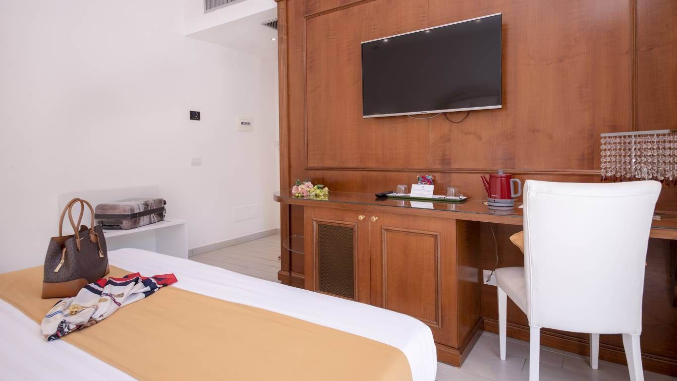A hotel room with a bed, a desk with a TV, a white chair, and a handbag on the bed.