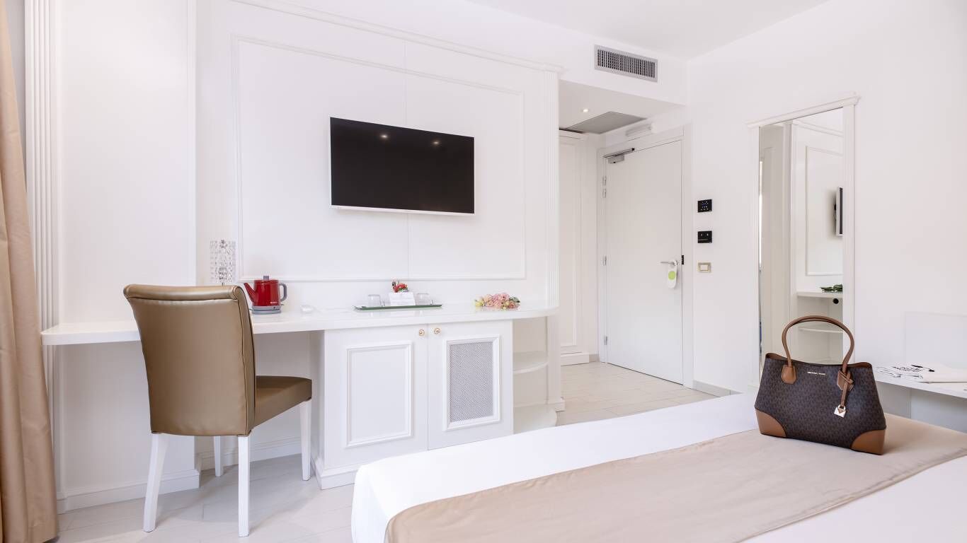 A hotel room with a bed, a desk with a chair, a TV mounted on the wall, and a handbag on the bed.