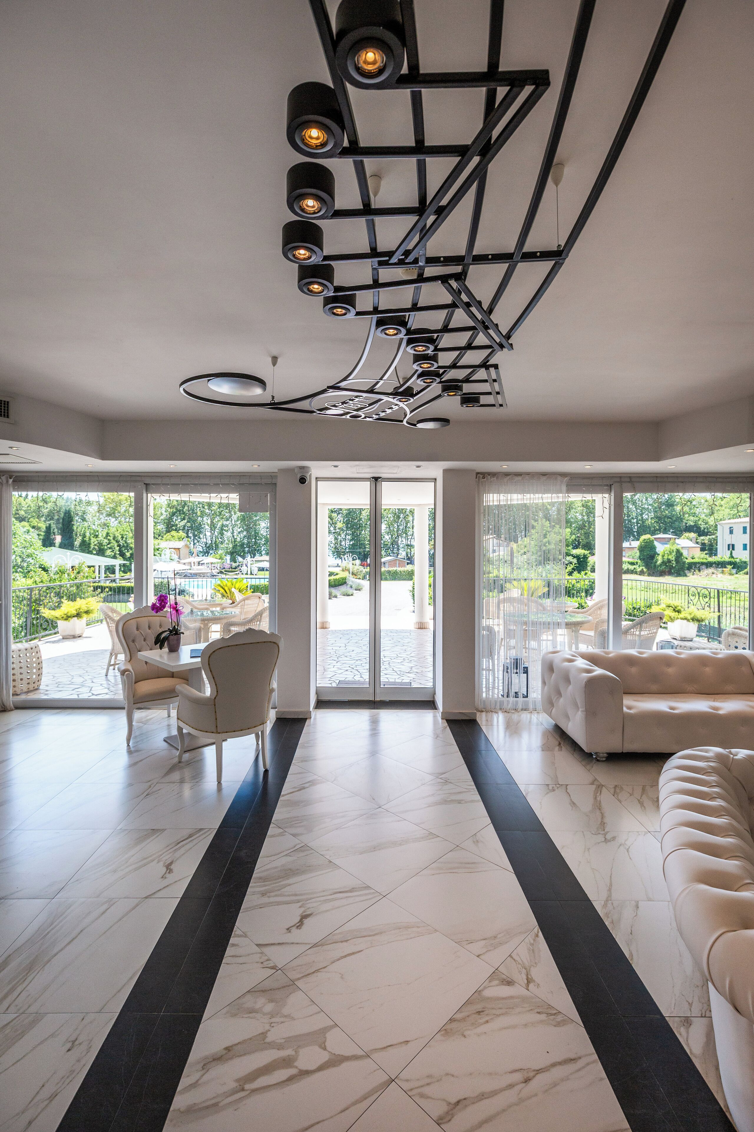 A spacious room with a large window, a white sofa, a dining table with chairs, and a modern ceiling light fixture.