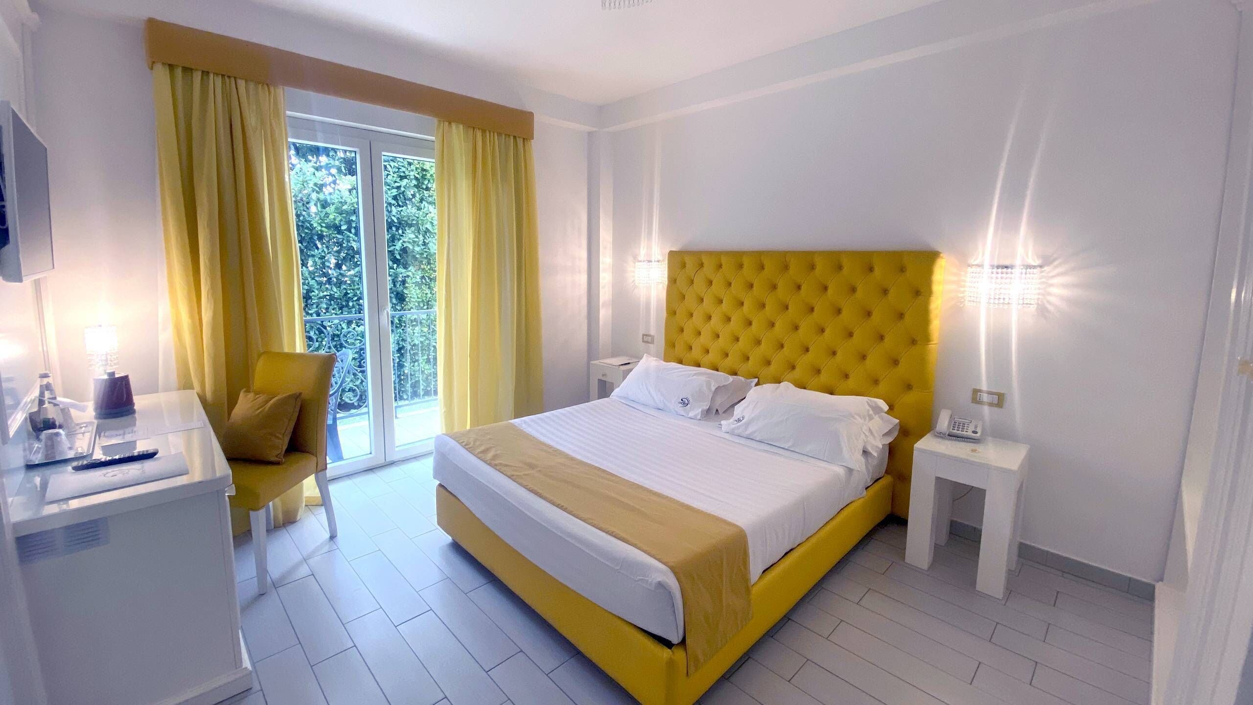 Quarto de hotel com uma cama grande, uma escrivaninha com cadeira, mesa de cabeceira e vista para o verde pela janela.