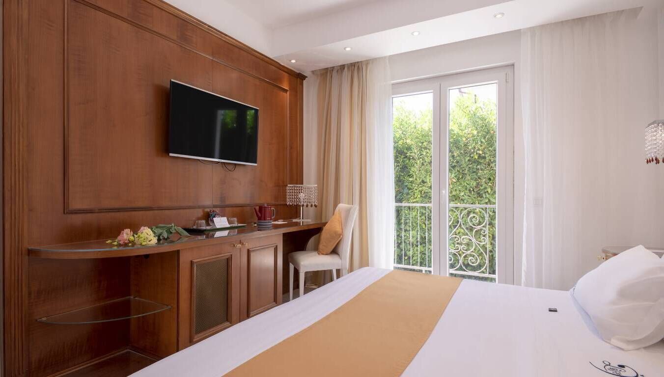 Habitación de hotel con una cama grande, un televisor montado en la pared, un escritorio con una silla y un balcón con vistas a zonas verdes.