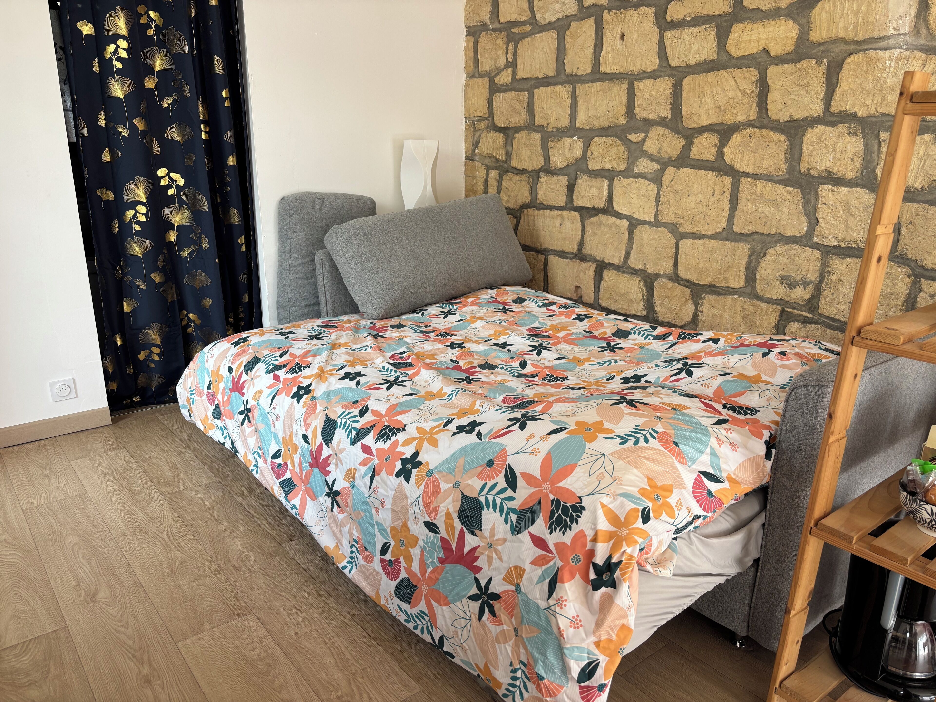 A bedroom with a floral bedspread, grey pillows, a stone wall, and a wooden side table with a kettle.