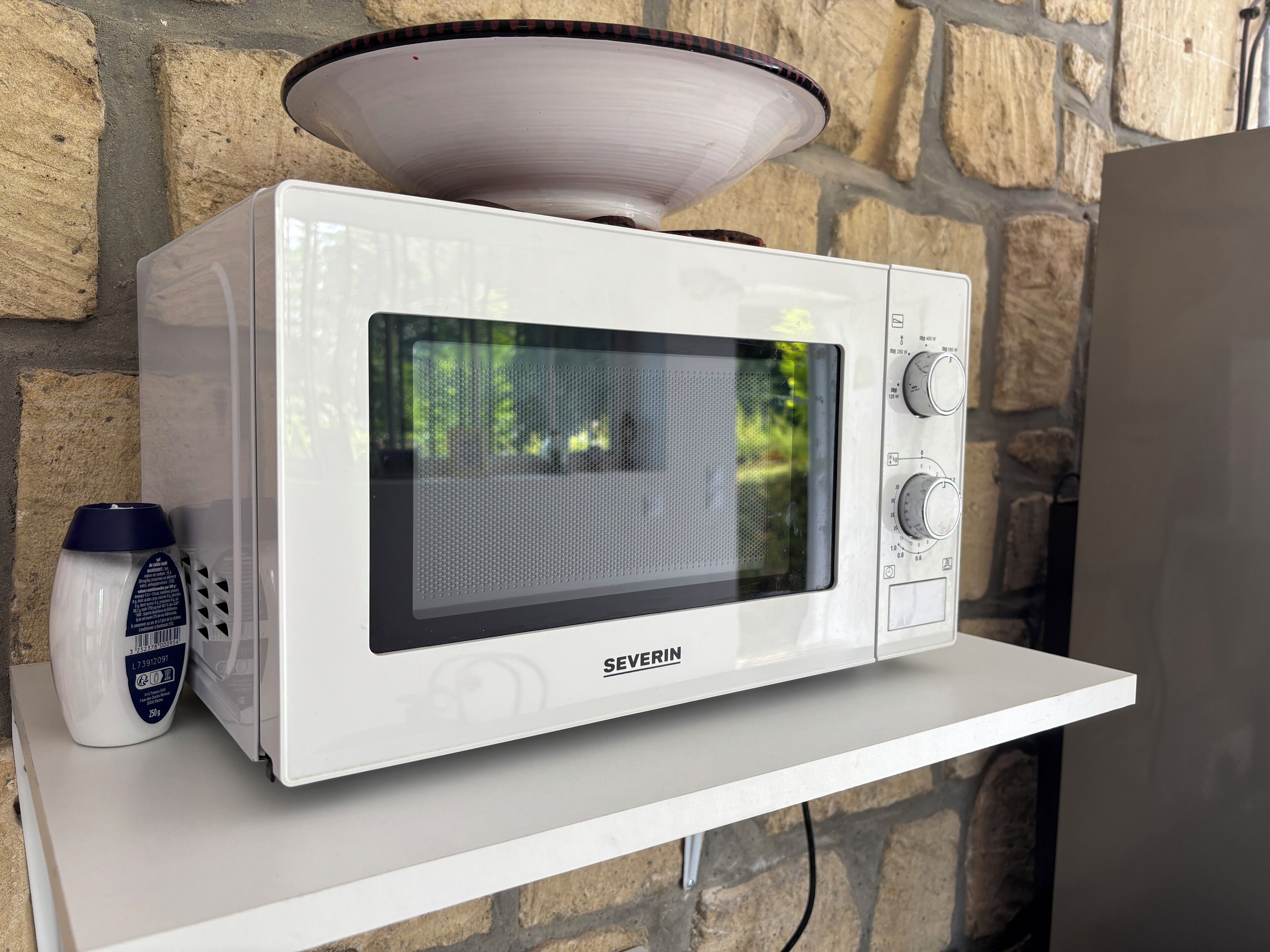 Un horno microondas blanco con pantalla digital y perillas de control, sobre una repisa blanca frente a una pared de piedra.