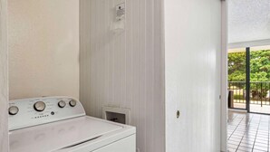 Une buanderie équipée d’un lave-linge blanc, d’un panneau de commande avec réglages de température et d’une porte donnant sur un balcon extérieur avec vue sur la verdure.