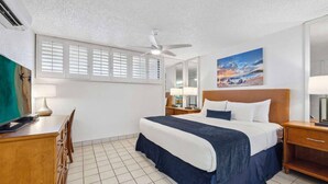 Ein Hotelzimmer mit einem Bett, einem Schreibtisch, einem Stuhl, einem Deckenventilator, einem Fenster mit Jalousien und einem Bild an der Wand.