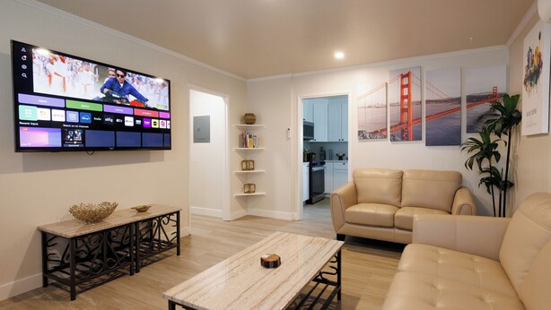 Un salon moderne avec un téléviseur à écran plat, un canapé beige, une table basse et une cuisine en arrière-plan.