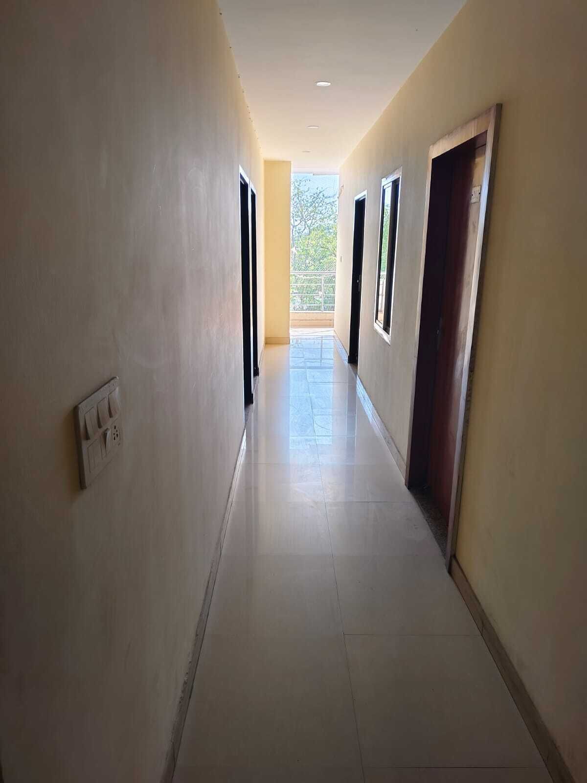 A long, narrow hallway with tiled floors and plain walls, leading to a balcony with a view of trees.