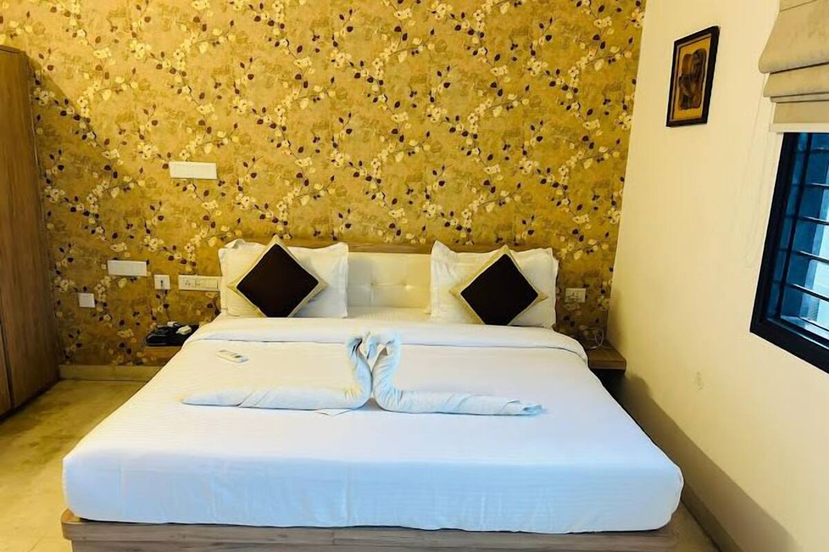 Quarto de hotel com papel de parede estampado com flores, uma cama com roupa de cama branca e duas almofadas, e uma cabeceira de madeira.
