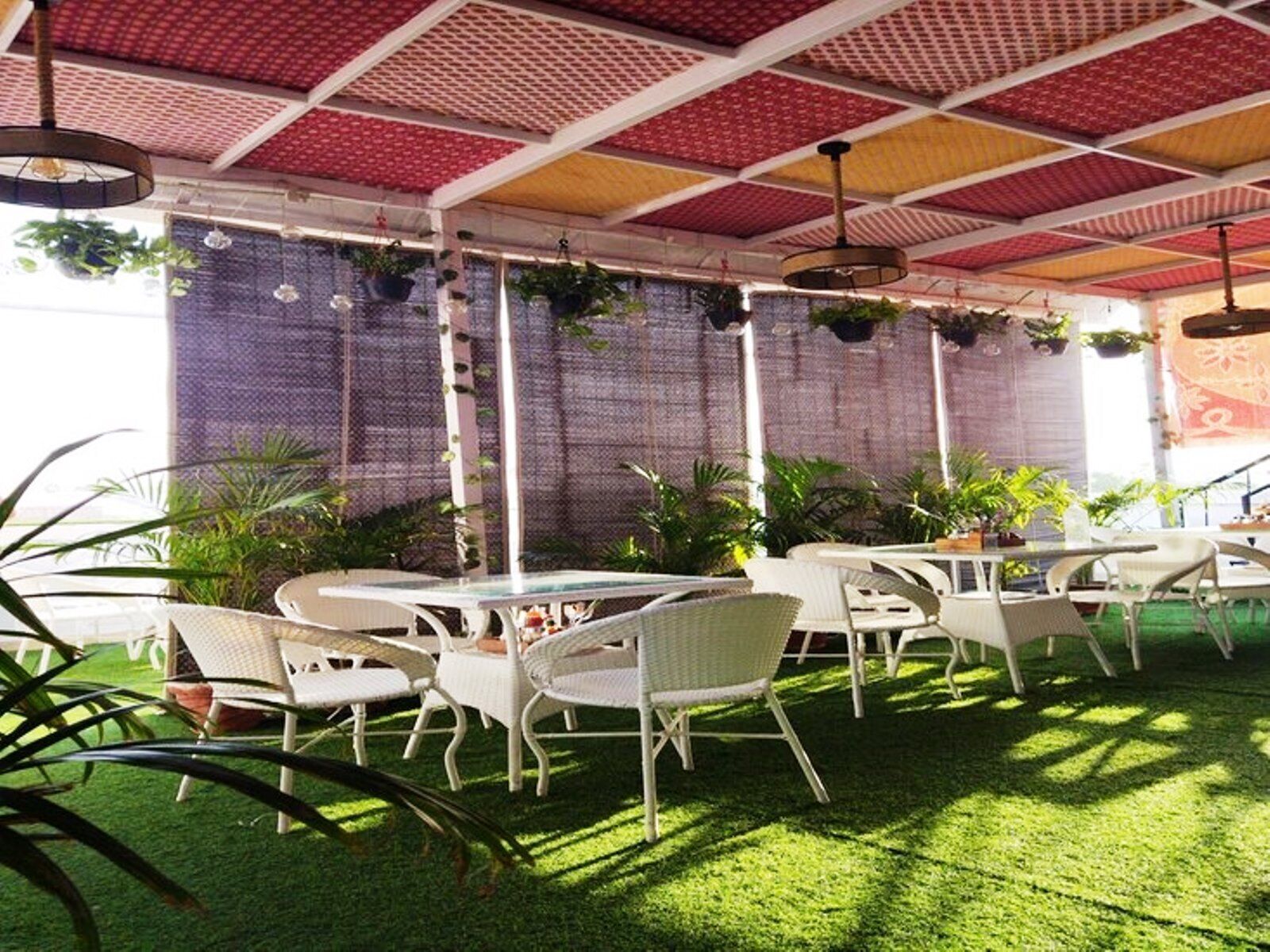 An outdoor seating area with round tables and white chairs, surrounded by green artificial grass, under a red and white patterned roof.