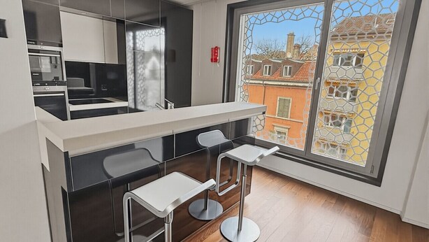 A modern kitchen with a built-in oven, a dining area with a white countertop and metal stools, and a large window with a view of buildings. - Comfortable 1BR flat / great location - Baron 44 (Zürich)