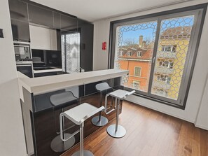A modern kitchen with a built-in oven, a dining area with a white countertop and metal stools, and a large window with a view of buildings. - Comfortable 1BR flat / great location - Baron 44 (Zürich)