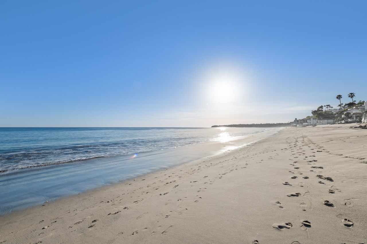 A beach with footprints leading to the water, a clear sky, and buildings in the background.