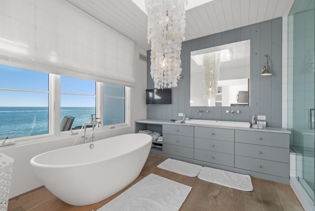 Un baño moderno con una bañera grande, muebles grises, un espejo amplio y vista al océano.