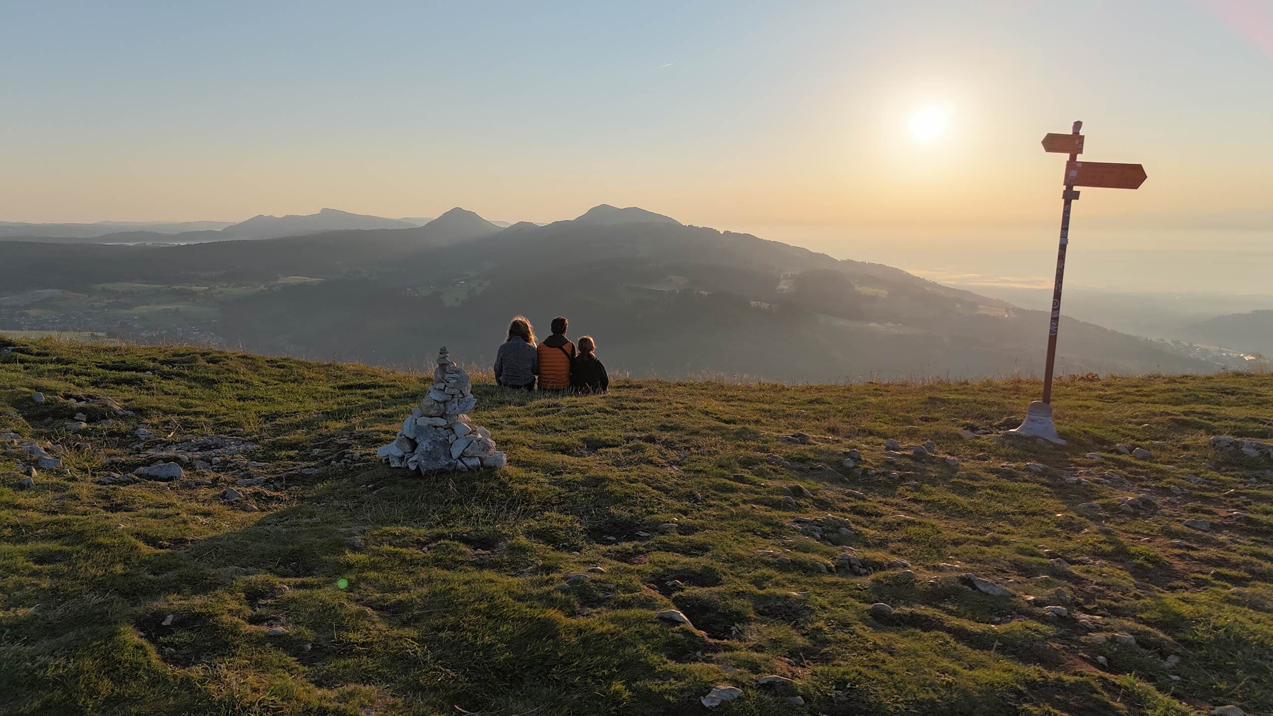 Una familia sentada en una colina cubierta de hierba, con vistas a una montaña y un cartel indicador.