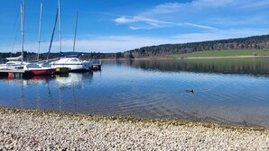 A calm lake with boats docked on the shore and a forested hill in the background.