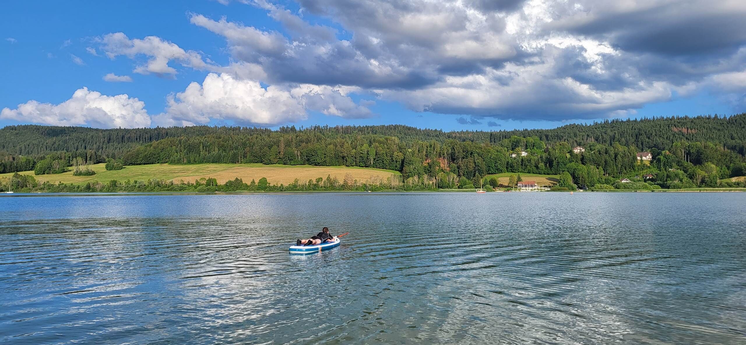 Un lac paisible où une personne se trouve sur un radeau bleu, entouré de collines verdoyantes et d’une zone boisée, sous un ciel partiellement nuageux.