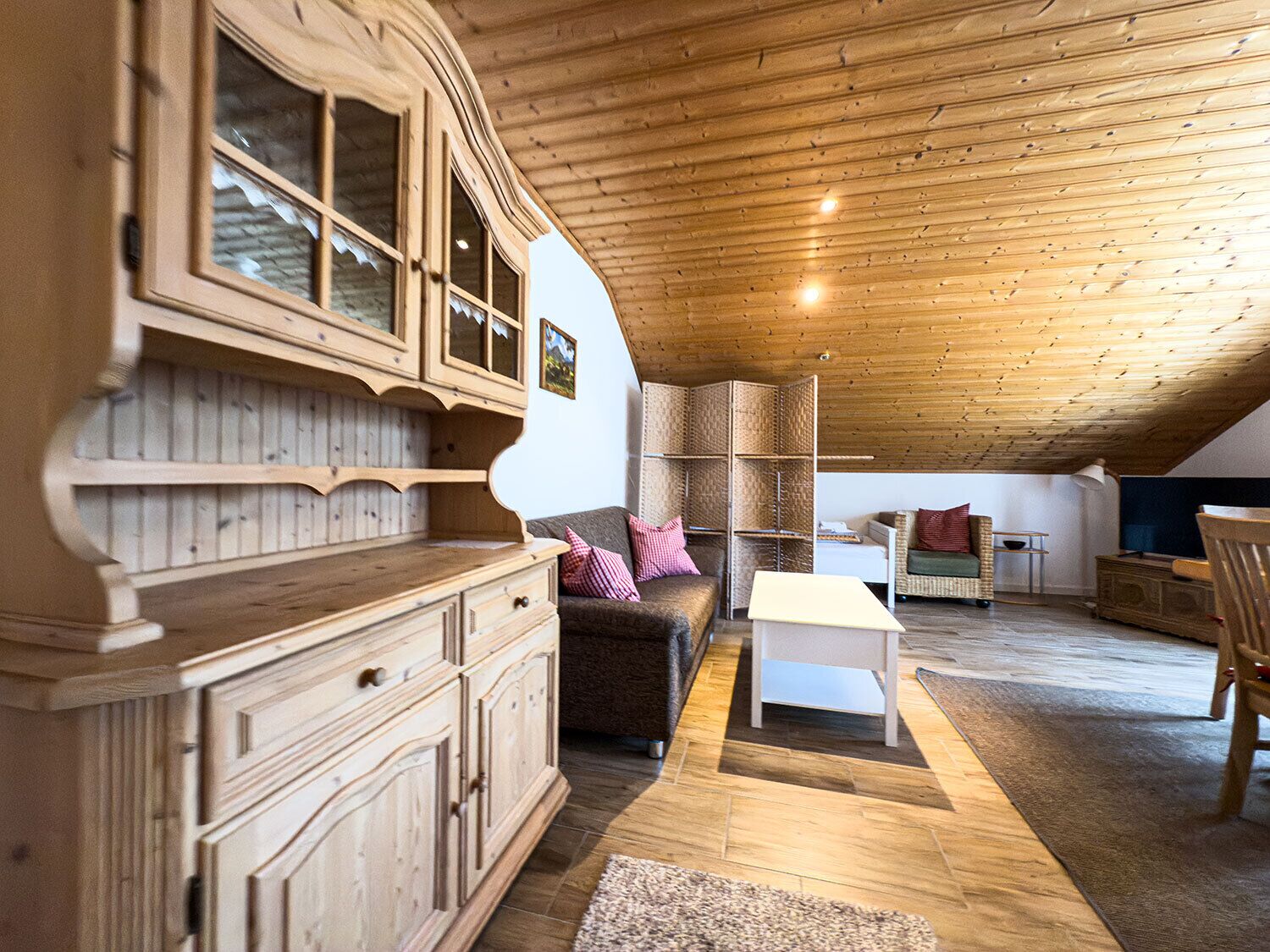 Ein gemütliches Dachzimmer mit Holzbalken und -wänden, möbliert mit einer Couch, einem kleinen Tisch und einem Schrank.