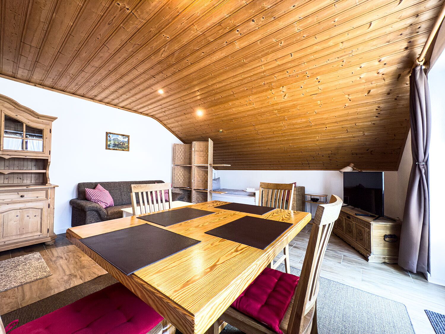 Ein gemütlicher, rustikaler Raum mit Holzbalkendecke, einem Esstisch mit schwarzen Platzsets, einer Couch mit rotem Kissen und einem Fernseher auf einem Holzschrank.