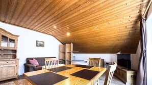 Ein gemütlicher, rustikaler Raum mit Holzbalkendecke, einem Esstisch mit schwarzen Platzsets, einer Couch mit rotem Kissen und einem Fernseher auf einem Holzschrank.