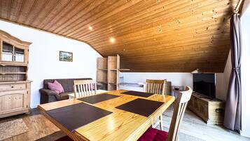 Ein gemütlicher, rustikaler Raum mit Holzbalkendecke, einem Esstisch mit schwarzen Platzsets, einer Couch mit rotem Kissen und einem Fernseher auf einem Holzschrank.