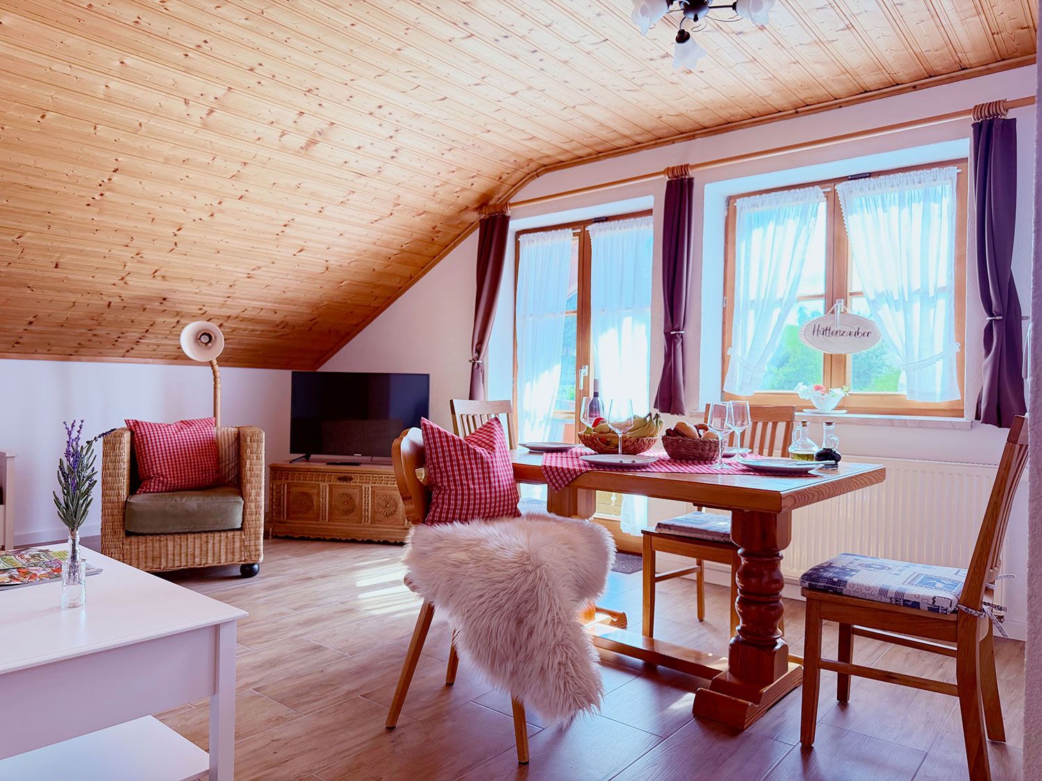 Une pièce chaleureuse avec un plafond en bois, une table à manger dressée pour deux personnes, un téléviseur et un fauteuil en osier.