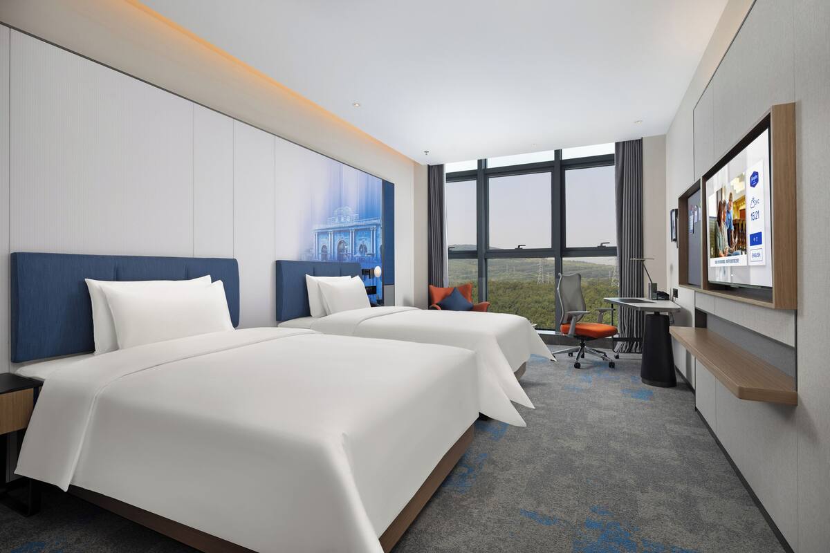 A hotel room with two beds, a desk, a chair, and a large window offering a view.