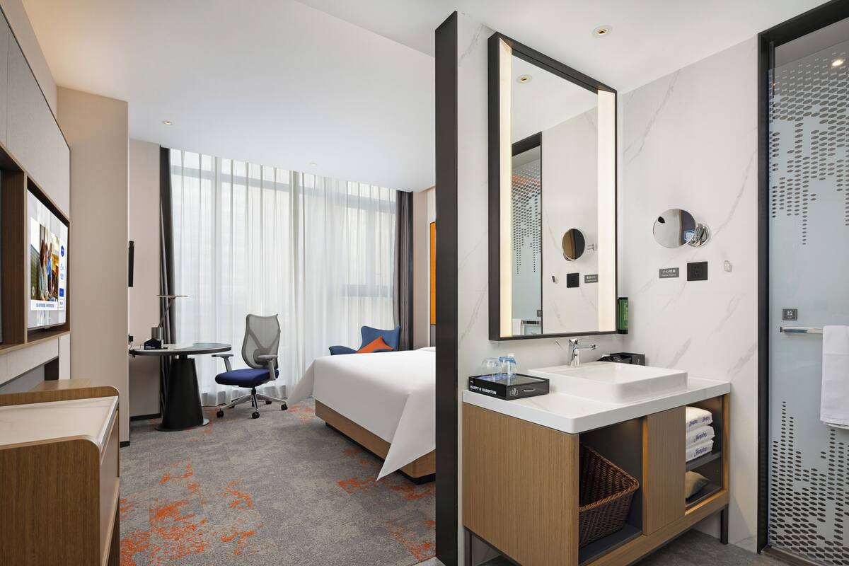 A hotel room with a bed, a desk, a chair, a large mirror, a bathroom with a sink, and a shower area.