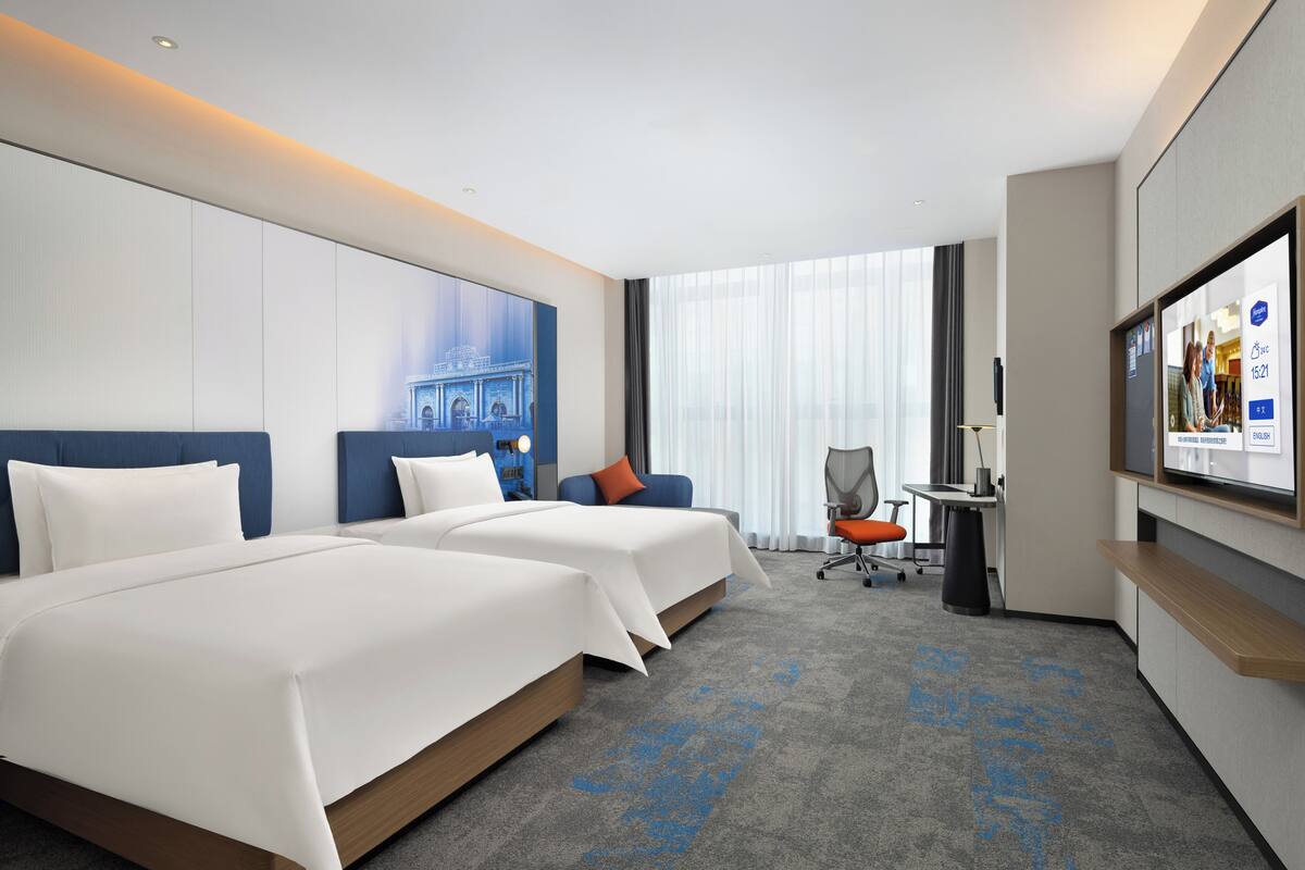A hotel room with two beds, a desk, a chair, a TV, and a large window with curtains.
