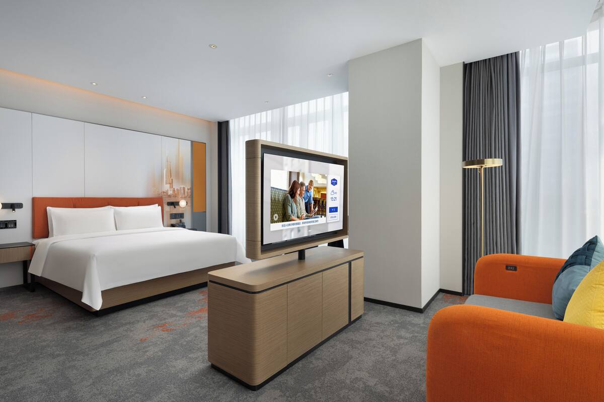 A modern hotel room with a bed, a TV on a stand, a desk, and a seating area.
