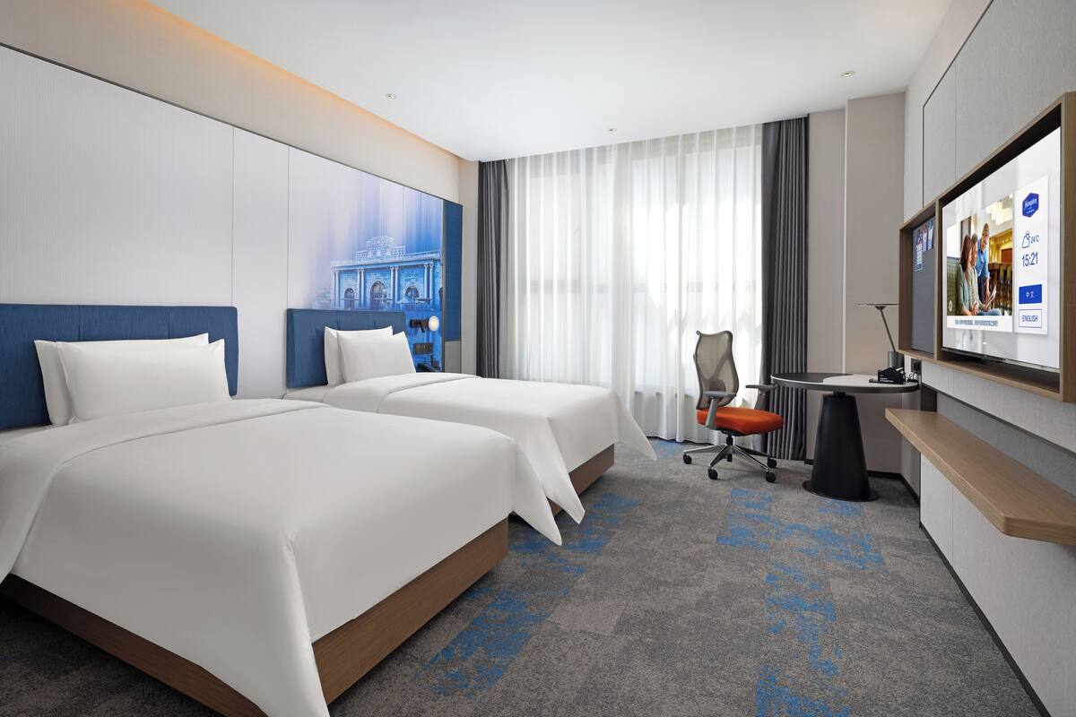 A hotel room with two beds, a desk, a chair, a TV, and a large window with curtains.