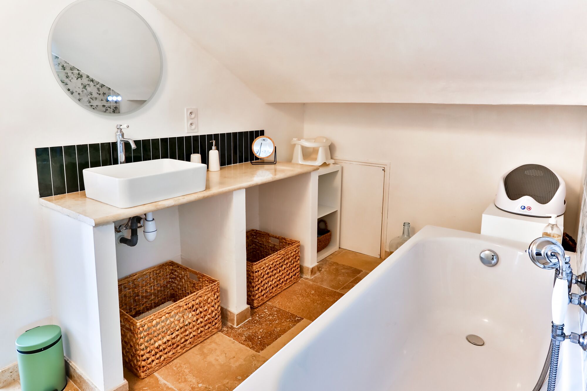 Ein Badezimmer mit weißem Waschbecken, einem runden Spiegel, einer Uhr, einem kleinen Ablagebereich für Toilettenartikel, einer Badewanne und Korbwannen.
