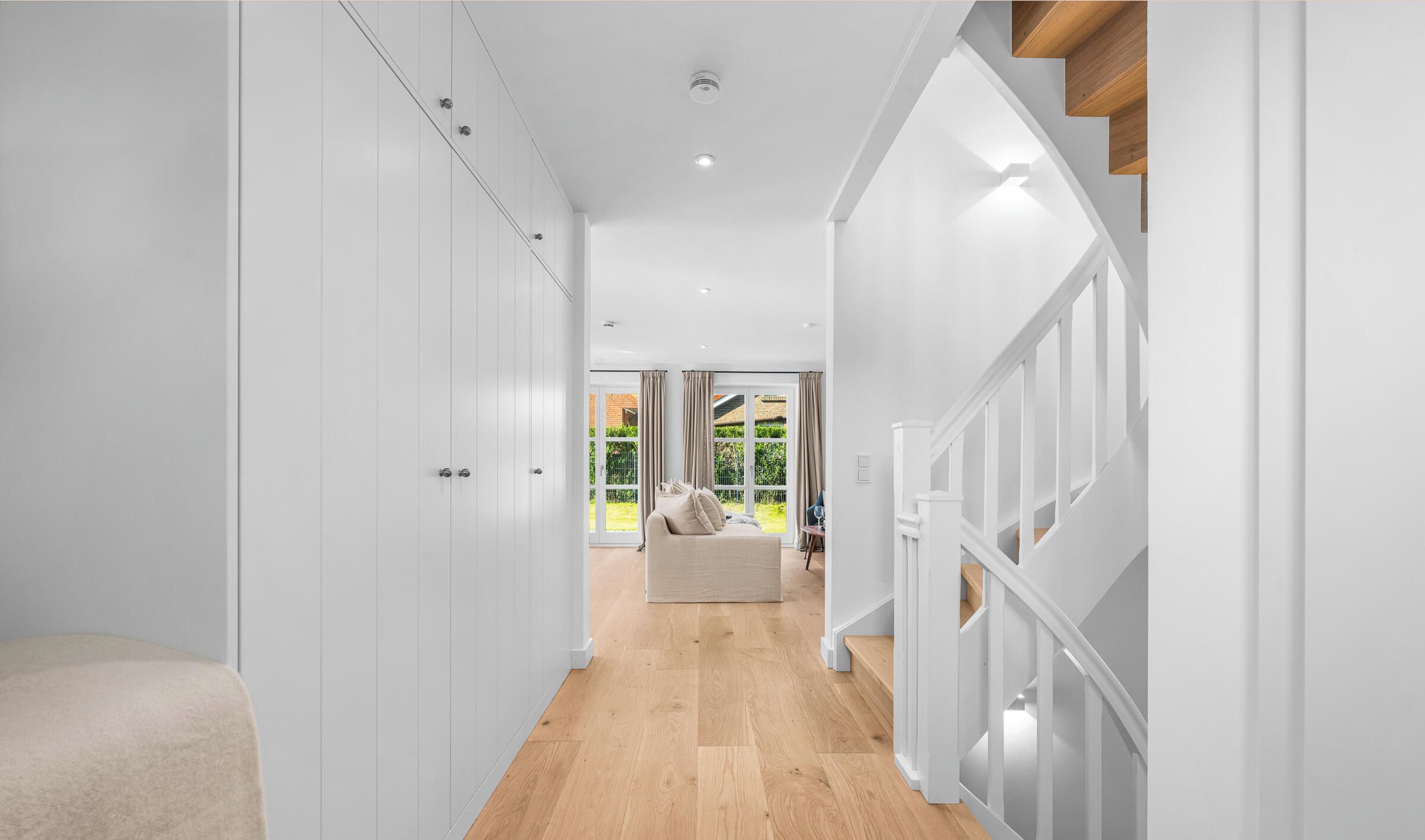 A hallway with wooden flooring, white walls, and a staircase with white railings.