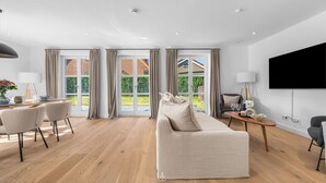 A modern living room with a sofa, dining table, and chairs, large windows with curtains, a flat-screen TV, and a wooden floor. - Spacious 5-Star Villa with Sauna, Garden and Private Terrace in Tinnum (Tinnum)