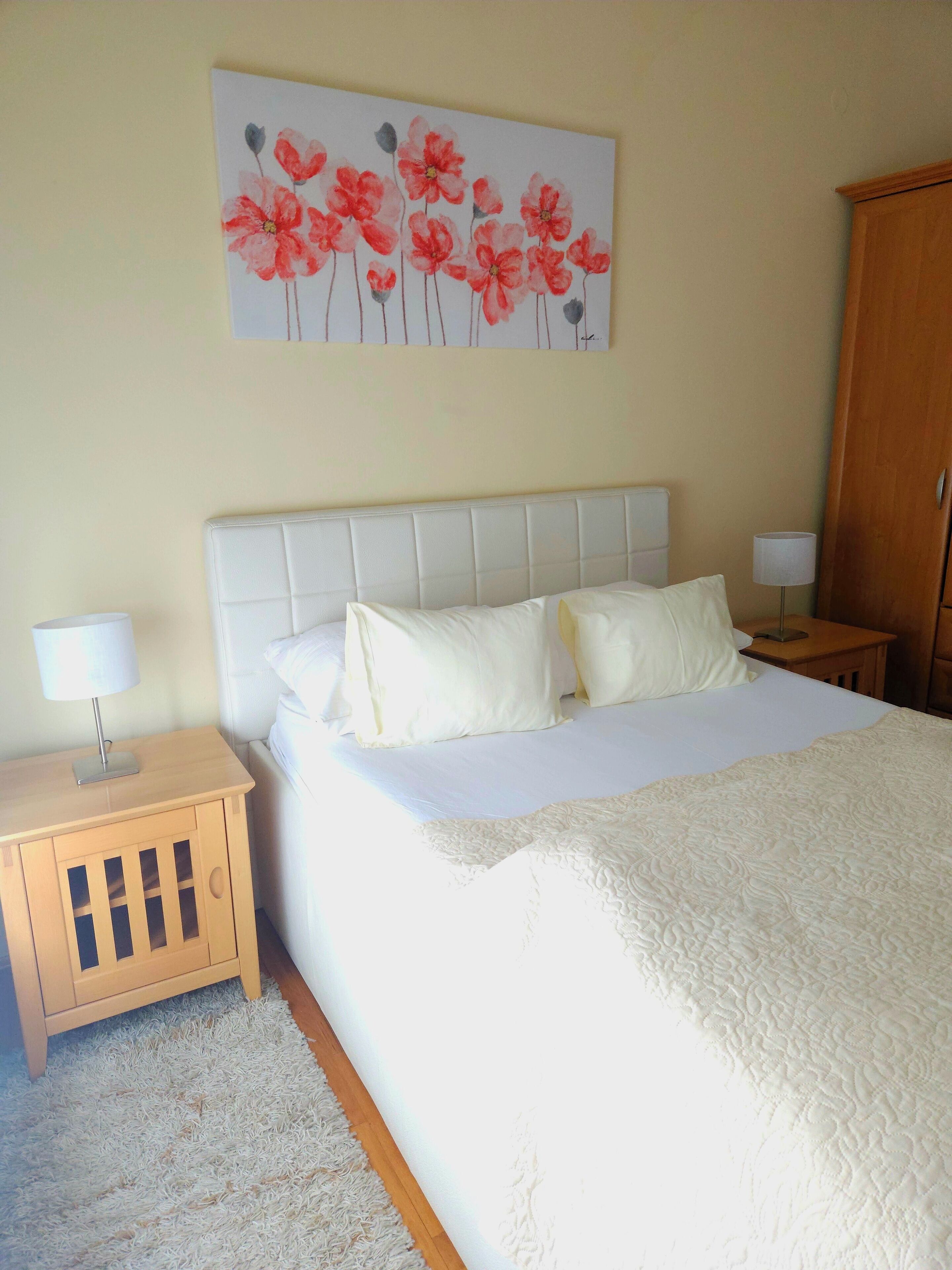 Une chambre à coucher avec un lit, deux tables de chevet, une armoire en bois et un tableau floral accroché au mur.
