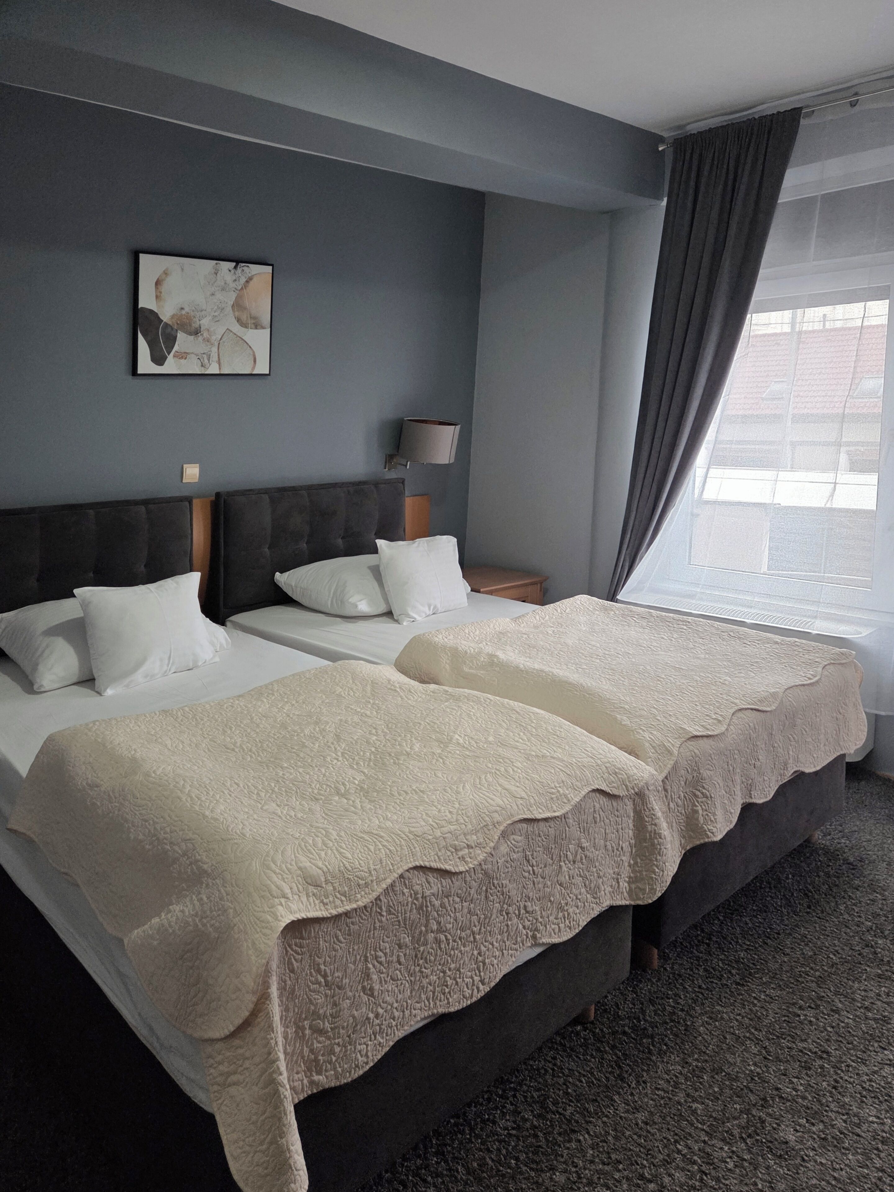 A hotel room with a large bed, two bedside tables, a lamp, a painting on the wall, and a window with curtains.