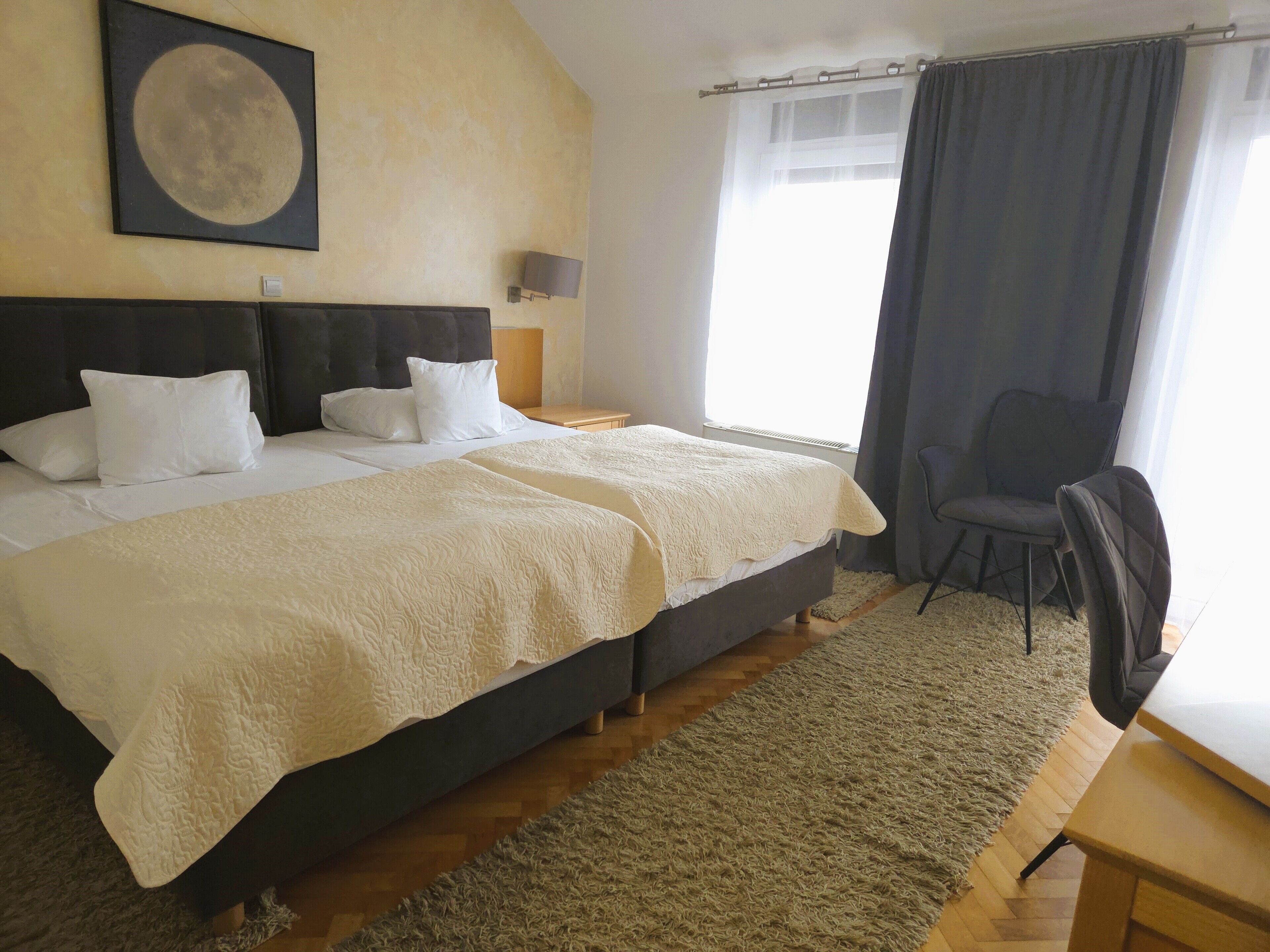 A bedroom with a bed, two pillows, a bedside table with a lamp, a chair, a large window with curtains, and a wall-mounted picture.