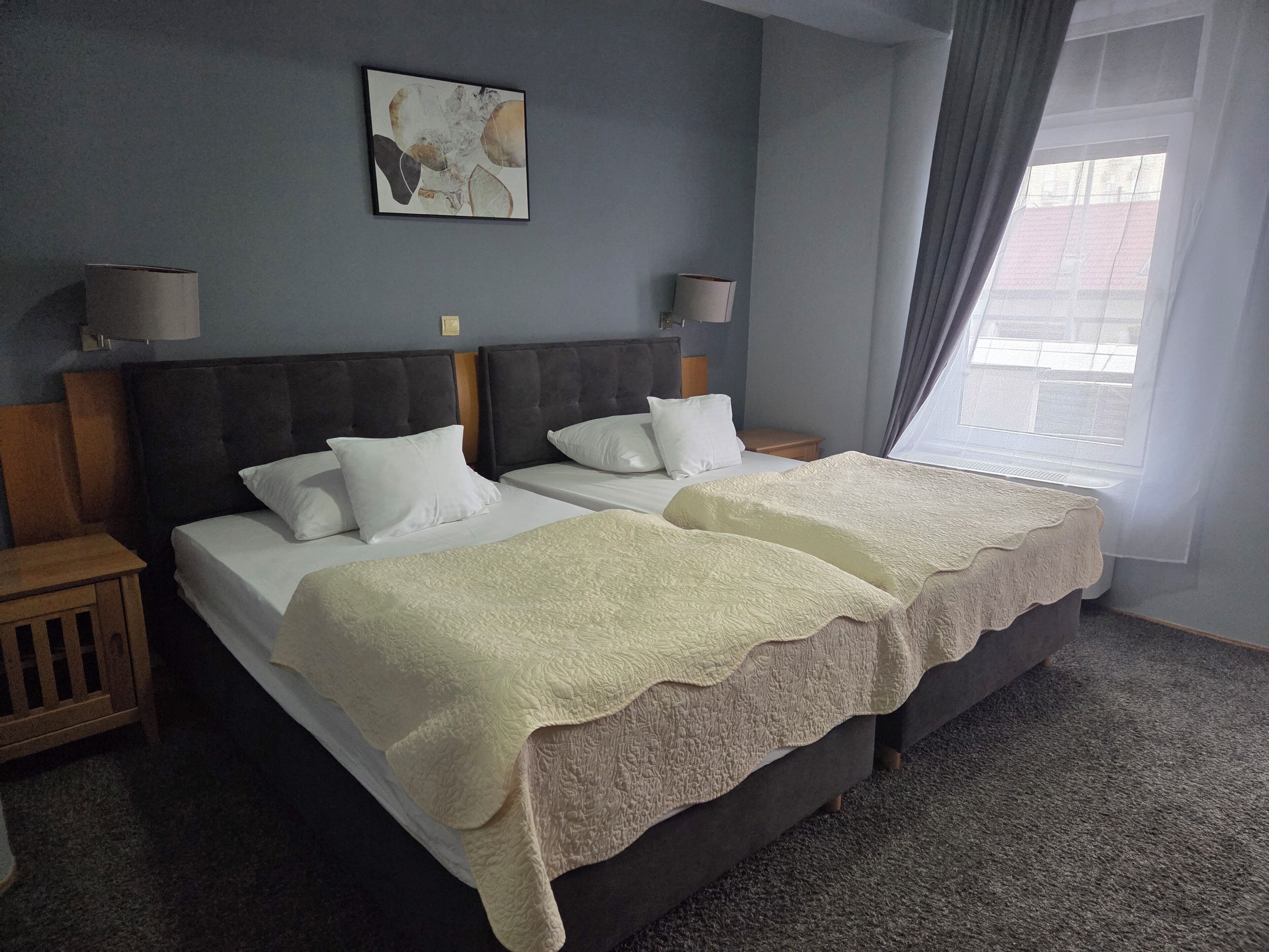 A double bed with two white pillows and a beige blanket, a bedside table with a lamp, a wall-mounted artwork, and a window with curtains.
