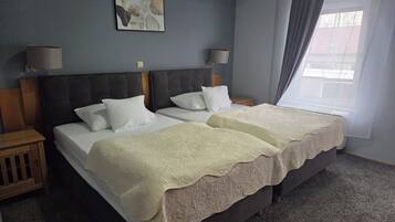 A double bed with two white pillows and a beige blanket, a bedside table with a lamp, a wall-mounted artwork, and a window with curtains.