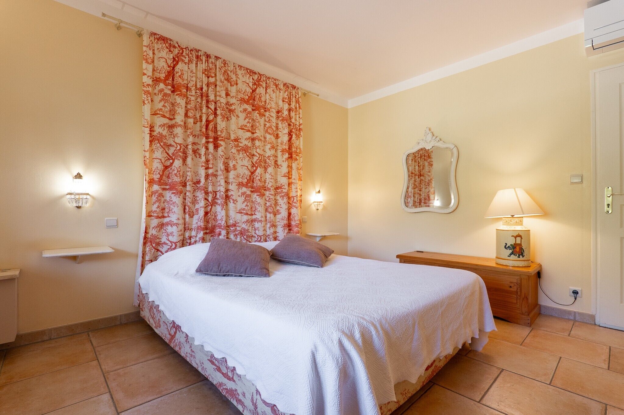 A bedroom with a bed, two bedside lamps, a nightstand with a lamp, a mirror, and a wall-mounted shelf.