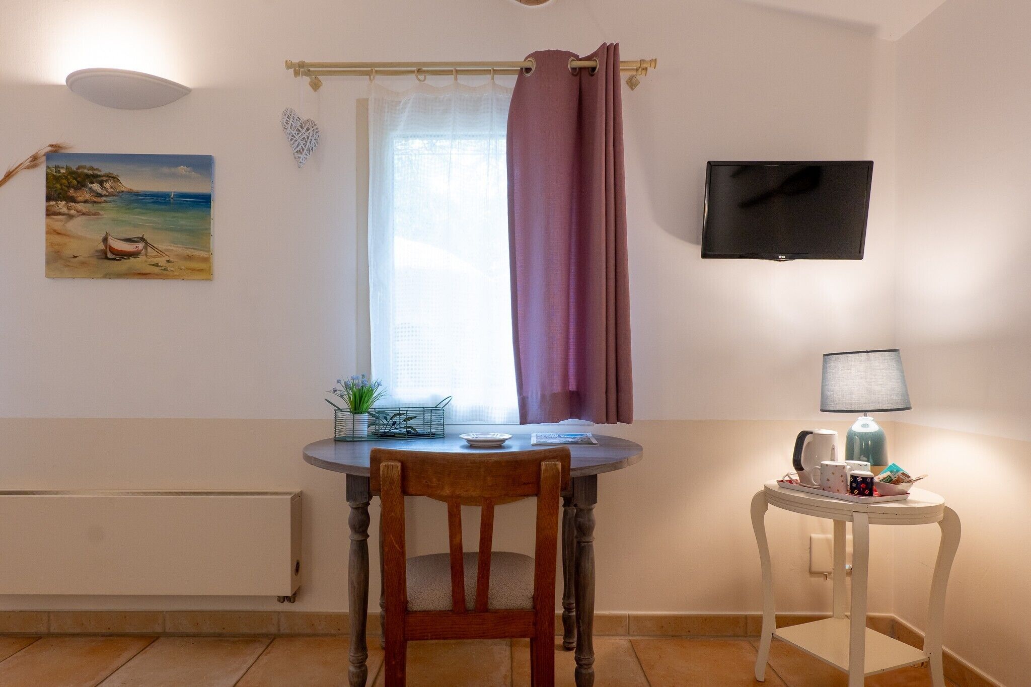 Ett rum med ett bord, en stol, en tv, en lampa och en tavla som föreställer en strandvy.