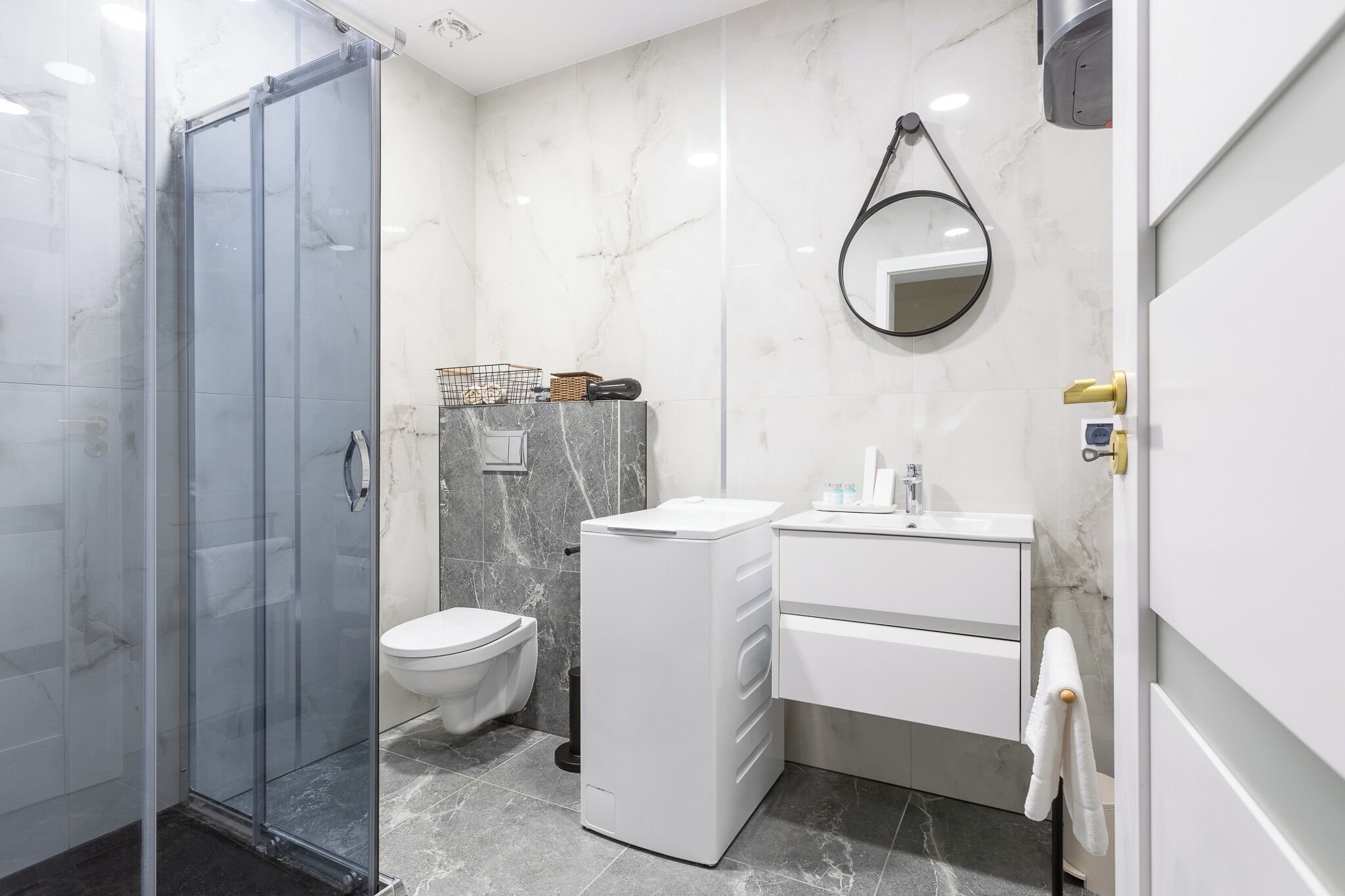 A modern bathroom with a glass shower enclosure, a toilet, a compact washing machine, and a sink with a mirror.