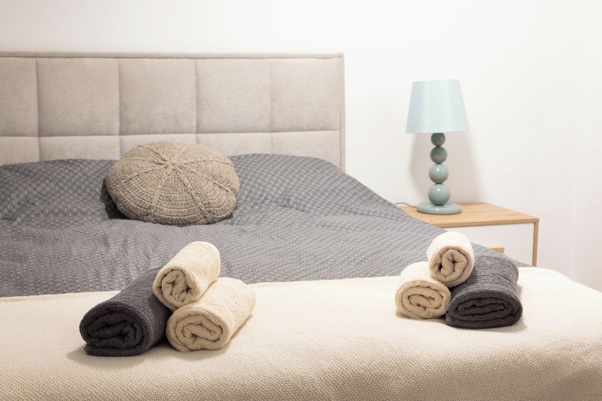 Una cama bien hecha con una colcha estampada en gris y beige, un almohadón gris y toallas enrolladas en tonos grises y beiges. Se ve una mesita de noche con una lámpara.