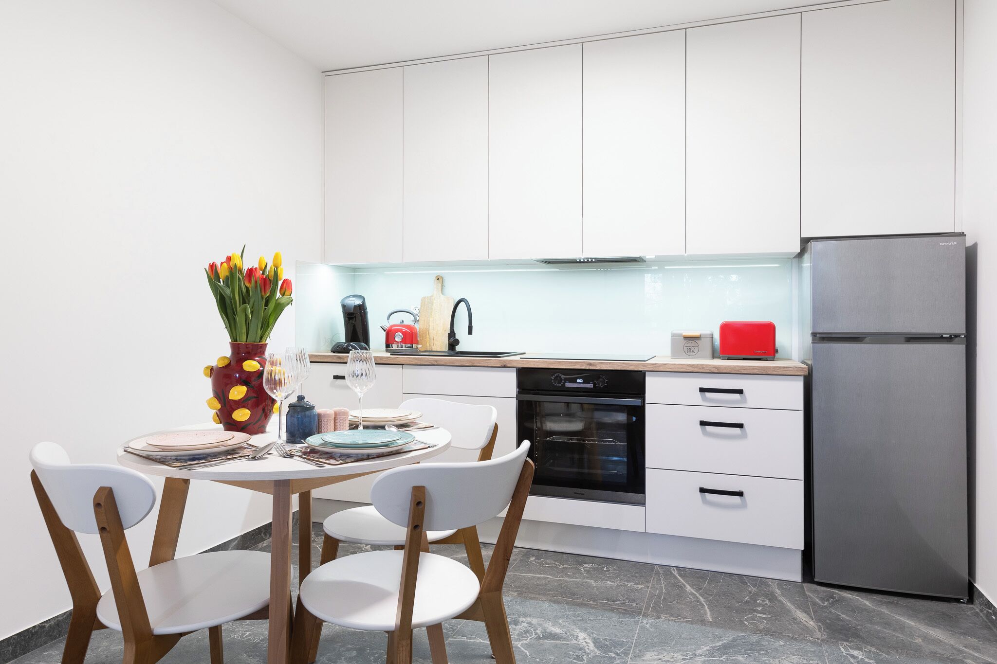 A modern kitchen with white cabinets, a stainless steel refrigerator, and a dining area with white chairs and a table set for two.