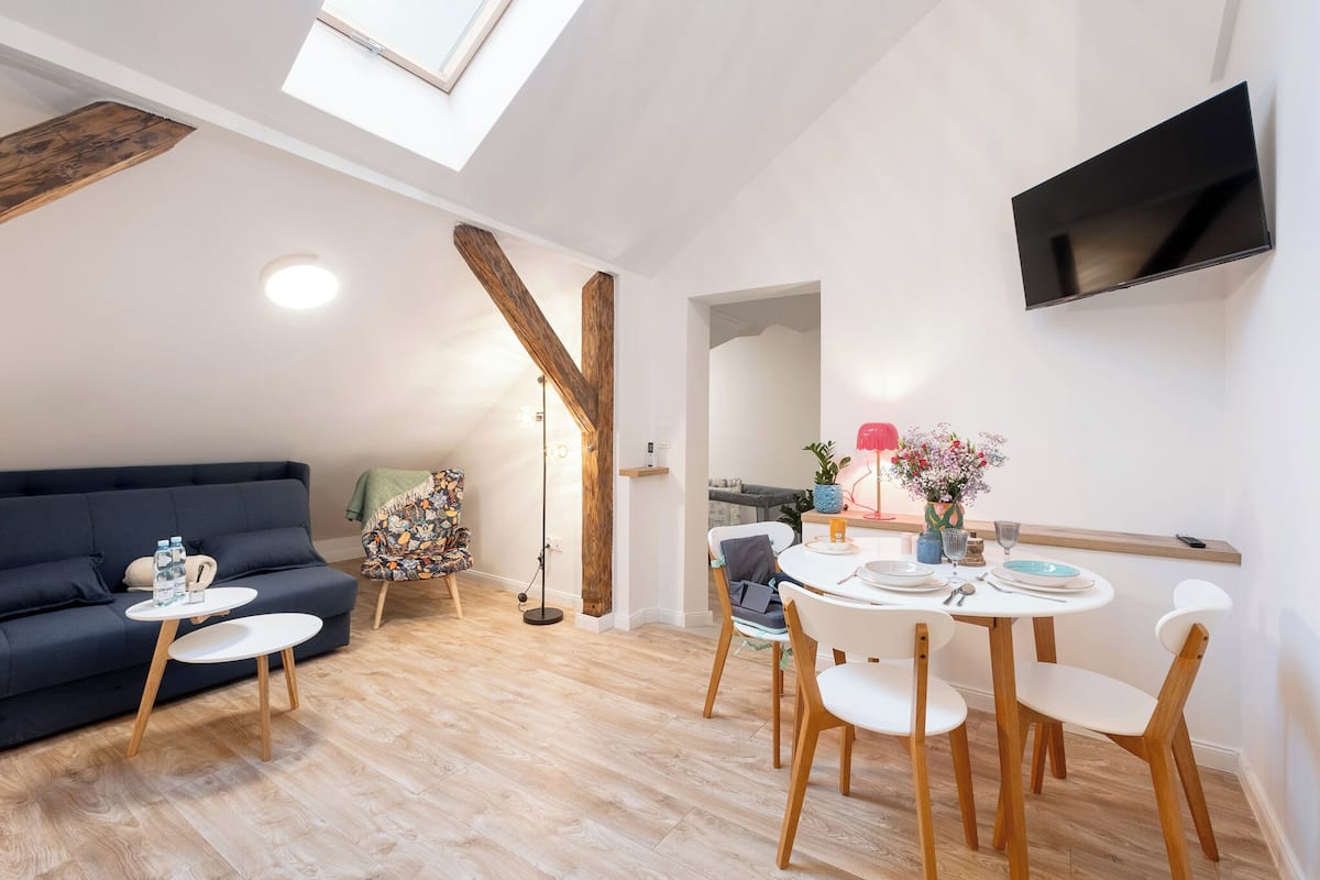 A modern living room with a sofa, two small tables, a dining table with chairs, a TV mounted on the wall, and a skylight.