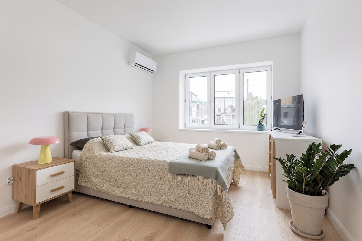 A bedroom with a bed, bedside table, TV, and a window with a view of buildings.