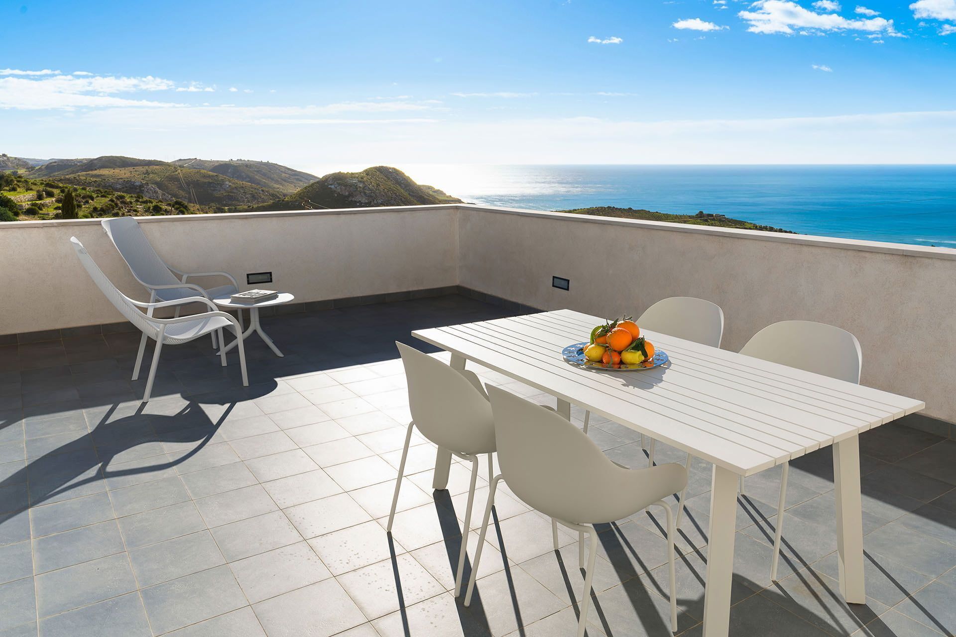 Eine Terrasse mit einem weißen Tisch und Stühlen, einer Obstschale und Blick auf das Meer und die Hügel.